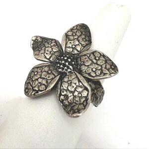 May include: A silver-toned ring featuring a large, five-petaled flower design. Each petal is embossed with a detailed pattern of smaller flowers and leaves. The center of the flower is textured with a cluster of small, raised dark dots.
