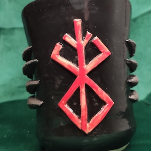 Puede incluir: Taza de cerámica negra con un símbolo rojo. La taza tiene forma cilíndrica con la parte superior ligeramente ensanchada y pequeñas protuberancias redondeadas en los lados. El símbolo es un diseño geométrico estilizado.