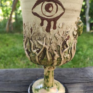 Op de afbeelding: Een keramieken kelk met een handgeschilderd bruin oog met druipende details bij de rand. Het lichaam is versierd met verhoogde, textuurrijke wortelachtige patronen in groen- en beigetinten en een kleine groene keversculptuur aan de zijkant.