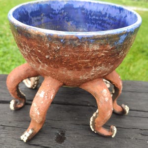 May include: A ceramic bowl with a textured, reddish-brown exterior and a smooth, deep blue interior. The bowl is supported by four curved, tentacle-shaped legs that feature small, white, circular suction cup details along their undersides.