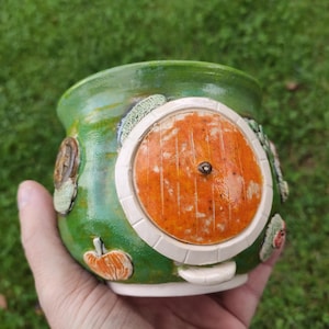 Pode incluir: Uma caneca de cerâmica verde com uma porta circular laranja em relevo com um puxador central, rodeada por uma moldura branca. O exterior é decorado com pequenos detalhes esculpidos, incluindo uma abóbora e folhagem, com acabamento brilhante.