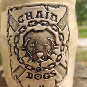 Może przedstawiać: Beżowy ceramiczny kubek z szczegółowym wytłoczonym wzorem. Wzór przedstawia głowę psa otoczoną łańcuchem, z napisami "CHAIN OF DOGS" i symbolem. Kubek ma lekko teksturowaną powierzchnię.