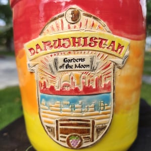 Może przedstawiać: Cylindryczny ceramiczny kubek z gradientem w kolorach czerwonym, pomarańczowym i żółtym. Wypukły emblemat zawiera napis "DARUSHISTAN" oraz "Gardens of the Moon" nad szczegółowym reliefem przedstawiającym panoramę miasta, mur i kiść winogron.