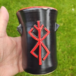 Könnte beinhalten: Schwarze Keramikbecher mit rotem Rand und rotem Symbol auf der Vorderseite. Der Becher hat zwei kleine, dreieckige Griffe. Das Symbol ist ein stilisiertes, geometrisches Design. Der Becher wird vor einem Hintergrund aus grünem Gras gehalten.