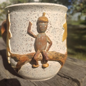 Pode incluir: Uma caneca de cerâmica com esmalte esbranquiçado salpicado, apresentando um personagem de bolota castanho esculpido em relevo sentado num ramo. O personagem tem uma cabeça redonda, membros finos e um rosto sorridente. Detalhes de folhas esculpidas.