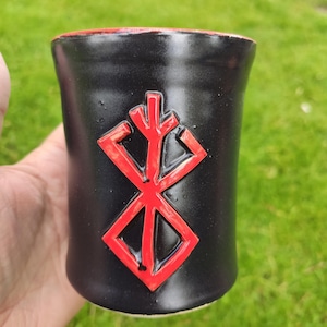 Op de afbeelding: Zwarte keramische mok met een rode binnenkant en een verhoogd rood symbool. De cilindrische mok lijkt handgemaakt. De achtergrond is wazig groen gras. De mok wordt vastgehouden.