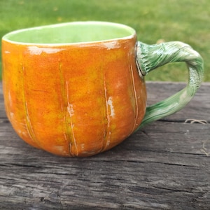 Peut inclure: Une tasse en céramique en forme de citrouille avec un extérieur orange brillant présentant des crêtes verticales et une anse verte texturée ressemblant à une tige. L'intérieur de la tasse est émaillé d'un vert clair contrastant avec le corps orange.