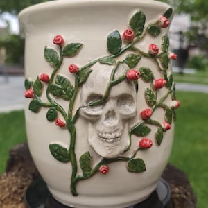 Puede incluir: Una taza de cerámica de color crema con un cráneo humano esculpido en tres dimensiones en el centro, rodeado de enredaderas verdes en relieve con pequeños capullos de rosa rojos. El diseño envuelve la superficie exterior del recipiente.