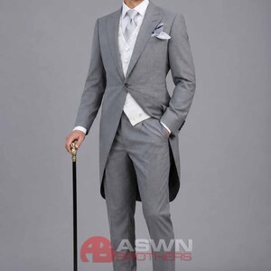May include: A grey morning suit featuring a long-tailed jacket, matching trousers, a white patterned waistcoat, a white dress shirt, and a light grey necktie. A pocket square is tucked into the jacket pocket, and a black cane with a gold handle is held.