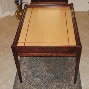 May include: A rectangular wooden table with a light-colored top and dark brown trim. The table has four legs and sits on a patterned rug. The top has a light wood center with a darker wood border and decorative lines.