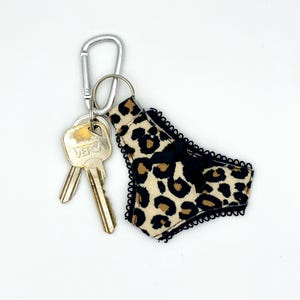 Può includere: Un portachiavi con una miniatura di biancheria intima in tessuto beige con stampa leopardata nera, bordo in pizzo nero e un piccolo fiocco nero al centro, fissato a un moschettone in metallo argentato con due chiavi in metallo.
