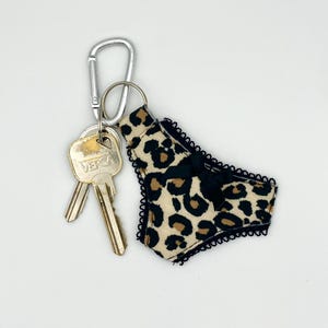 Può includere: Portachiavi con un ciondolo a forma di mutandine con stampa leopardata, bordatura in pizzo nero e fiocco nero. Il portachiavi include due chiavi e un moschettone argentato. Lo sfondo è bianco.