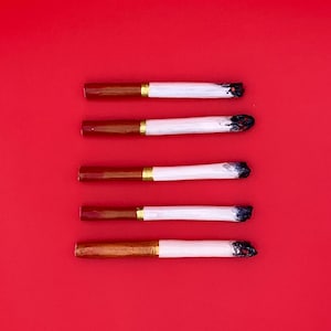 以下が含まれることがあります： 茶色のフィルター、白い紙、黒い灰の先端が付いた5本のハンドメイドタバコが、無地の赤い背景に対して垂直に配置されています。各タバコには、フィルターの近くに金のバンドが付いています。真上からの眺め。