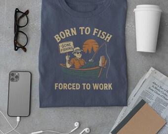 May include: A navy blue t-shirt with the text "BORN TO FISH, FORCED TO WORK" and a graphic of a person fishing. The image includes a smartphone, glasses, a coffee cup, and a newspaper.
