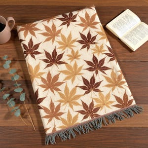 May include: A folded rectangular throw blanket with a cream background and a pattern of brown and tan maple leaves. A cup of coffee and an open book are next to the blanket. A sprig of eucalyptus is also present.