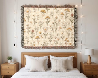 Könnte beinhalten: Eine rechteckige gewebte Wurfadecke mit einem Blumenmuster aus Gänseblümchen und anderen Wildblumen in Creme-, Gelb- und Brauntönen. Die Decke hat einen Fransenrand und wird über einem Bett mit weißen Kissen und einem hölzernen Kopfteil ausgestellt.