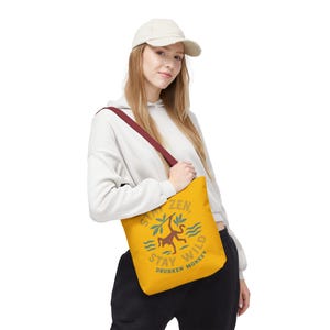 Puede incluir: Bolso tote mostaza con una correa granate. El bolso presenta un gráfico de un mono, hojas de palma y las palabras "Stay Zen, Stay Wild, Drunken Monkey" en un estilo vintage. El bolso se lleva al hombro.
