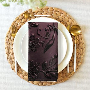 May include: A folded, deep purple napkin with embossed floral and leaf patterns, placed on a white plate. The plate is set on a woven placemat, with gold-coloured cutlery. The napkin's design features detailed flowers and foliage.