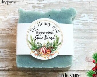 May include: A rectangular sage green bar of soap wrapped in a white paper band. The label features the text The Honey Well Peppermint Spice Blend, surrounded by a circular illustration of pine branches, red berries, and a striped ornament.