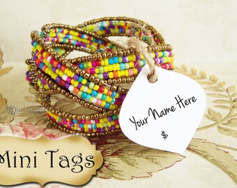 Peut inclure: Un bracelet en perles colorées avec une étiquette blanche sur laquelle est inscrit "Your Name Here". L'étiquette est attachée au bracelet avec de la ficelle. Le bracelet est sur un fond à motifs floraux.