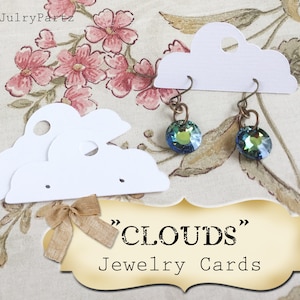 Op de afbeelding: Twee witte, wolkvormige sieraden displaykaarten met gaten om oorbellen op te hangen. De kaarten liggen op een bloemige achtergrond met een bord waarop "CLOUDS" Sieradenkaarten staat.