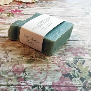 Puede incluir: Una pastilla de jab&oacute;n verde con una etiqueta blanca que dice "Soul Feather Studio" y enumera los ingredientes: "Ingredientes: aceite de oliva, aceite de coco, aceite de ricino, con hierbas infusionadas en aceite, algas verdes y aceites esenciales".