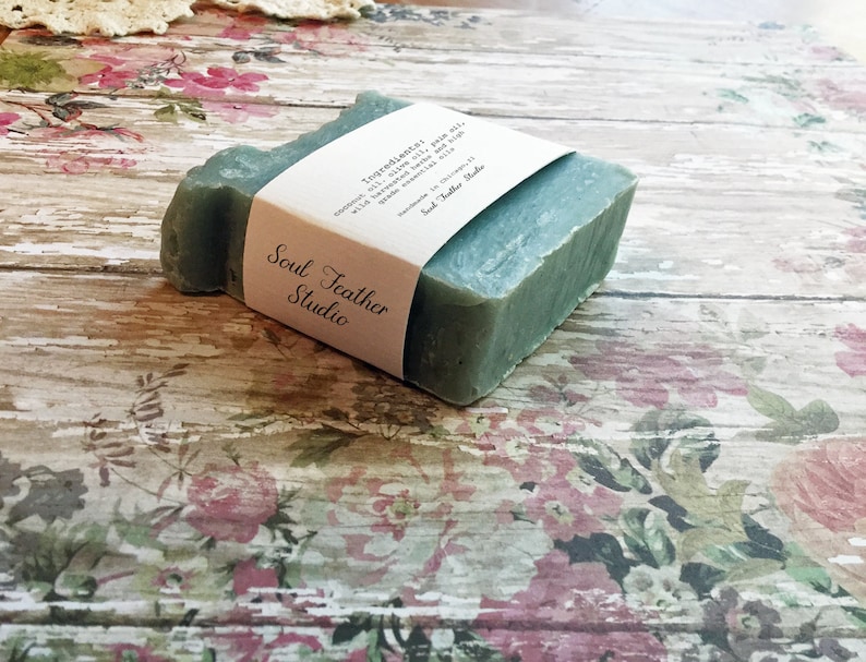 Puede incluir: Una pastilla de jab&oacute;n verde con una etiqueta blanca que dice "Soul Feather Studio" y enumera los ingredientes: "Ingredientes: aceite de oliva, aceite de coco, aceite de ricino, con hierbas infusionadas en aceite, algas verdes y aceites esenciales".
