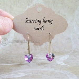 May include: A pair of gold hoop earrings with a purple heart-shaped crystal dangle. The earrings are hanging on a brown card that says "Earring hang cards".