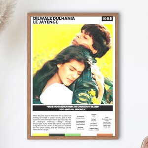 May include: A framed movie poster for "Dilwale Dulhania Le Jayenge" from 1995. The poster features a couple embracing, with text at the top and bottom. The frame is brown, and the poster is hanging on a white wall.