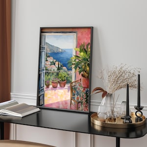 Puede incluir: Una pintura de acuarela enmarcada representa un balcón con vistas a un pueblo costero y al mar. La pintura presenta plantas en macetas, una silla pequeña y una vibrante paleta de colores rosa, azul y verde. La obra de arte se exhibe sobre una mesa negra con objetos decorativos.