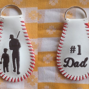 May include: Two white leather baseball-shaped keychains with red stitching. One keychain features a silhouette of a father and son playing baseball. The other keychain has the text "#1 Dad" embroidered in black.