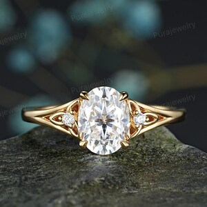 Puede incluir: Un anillo de oro con una gran piedra central blanca de corte ovalado sujeta por cuatro garras. La banda tiene un diseño de vástago dividido con una pequeña piedra blanca redonda engastada a cada lado dentro de un marco de metal decorativo.