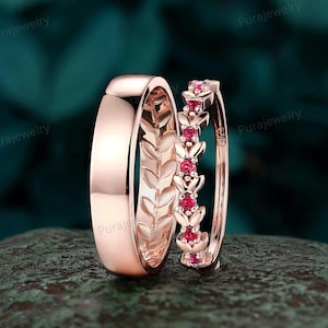 May include: Rose gold wedding ring set. One ring features a smooth, polished band, while the other has a delicate design with small red gemstones. The rings are displayed on a textured, dark green surface.