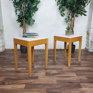 May include: Two wooden side tables with square white tops and light brown legs. The tables are placed on a wood-effect floor. One table has books on top, the other has a small potted plant. Tall green plants are in the background.