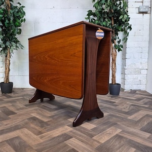 May include: A mid-century modern drop-leaf table featuring a warm, honey-toned wood grain surface on the leaves and a darker, rich walnut-coloured base with curved, sculptural legs. The table is shown in a folded position with a small circular tag.