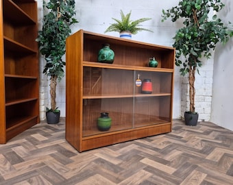 May include: A mid-century modern wooden bookcase with glass sliding doors. The bookcase has multiple shelves and is filled with decorative items. The wood has a warm, reddish-brown tone. The background includes potted plants and a white wall.