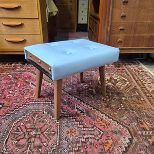 May include: A light blue upholstered footstool featuring a tufted top with four buttons, a dark wood frame with visible metal hardware on the side, and four tapered wooden legs. The rectangular seat is padded and sits atop the four legs.