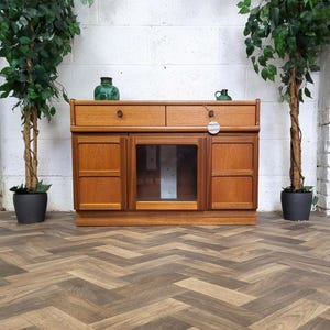 May include: A mid-century style wooden cabinet with two drawers and two doors, with a glass-fronted central section. The cabinet is a warm brown colour. Two green vases are placed on top.