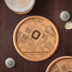 Könnte beinhalten: Ein runder Holzuntersetzer mit einer detaillierten Karte von Mönchengladbach, Deutschland, und seinen Koordinaten. Der Untersetzer ist hellbraun mit einer dunkelbraunen Stadiongrafik. Zwei Kronkorken und ein Bierglas sind ebenfalls zu sehen.
