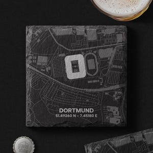 Könnte beinhalten: Ein quadratischer Untersetzer aus schwarzem Schiefer mit einer eingravierten Karte von Dortmund, Deutschland, wobei das Stadion weiß hervorgehoben ist. Der Text DORTMUND und die Koordinaten 51.49260 N - 7.45180 E sind unten in den Stein eingraviert.