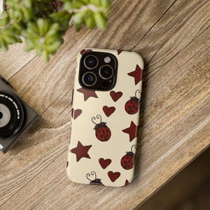May include: A cream-colored phone case featuring a pattern of red ladybugs, stars, and hearts. The ladybugs have black heads and red spotted bodies. The stars and hearts have a swirling design. The case has a camera cutout.