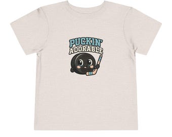 May include: A light beige toddler t-shirt featuring a cartoon hockey puck with a smiling face and a hockey stick. The text "PUCKIN' ADORABLE" is above the puck in blue and black letters.