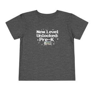 May include: Dark gray toddler t-shirt with the text "New Level Unlocked: *Pre-K*" in white pixelated font. A colorful pixelated video game controller graphic is below the text. The shirt is made of a soft material.
