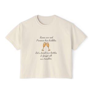 Op de afbeelding: Crèmekleurig T-shirt met de tekst "Roses are red, Prosecco has bubbles. Let's drink two bottles & forget all our troubles." Twee champagneglazen zijn eronder afgebeeld. Een casual kledingstuk.