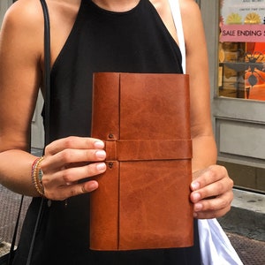 Peut inclure: Un journal en cuir marron avec une fermeture à lanière est présenté. Le journal est de forme rectangulaire et a une surface texturée. En arrière-plan, une boutique avec une enseigne "WAREHOUSE SALE UP TO 50% OFF".