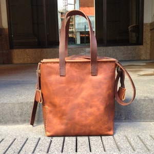 May include: A brown leather tote bag with a top zipper closure and a long strap. The bag has a distressed look and is made of high-quality leather.