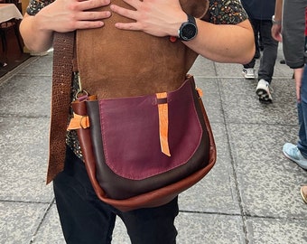 May include: A person wearing a black and brown leather satchel bag with a burgundy flap and orange accents. The bag has a long strap and is worn over the shoulder.