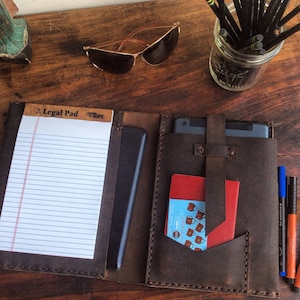 May include: A brown leather portfolio with a legal pad, a tablet sleeve, and a pocket for a small notebook. The portfolio has stitching around the edges and a flap with a strap.