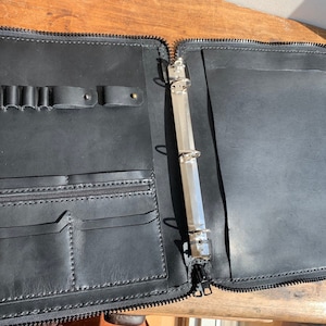 May include: Black leather zippered binder with a silver ring mechanism. The binder has a pocket on the front and a pencil holder on the top.