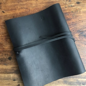 Peut inclure: Un journal en cuir noir avec une fermeture à boucle. Le journal est posé à plat sur une surface en bois.